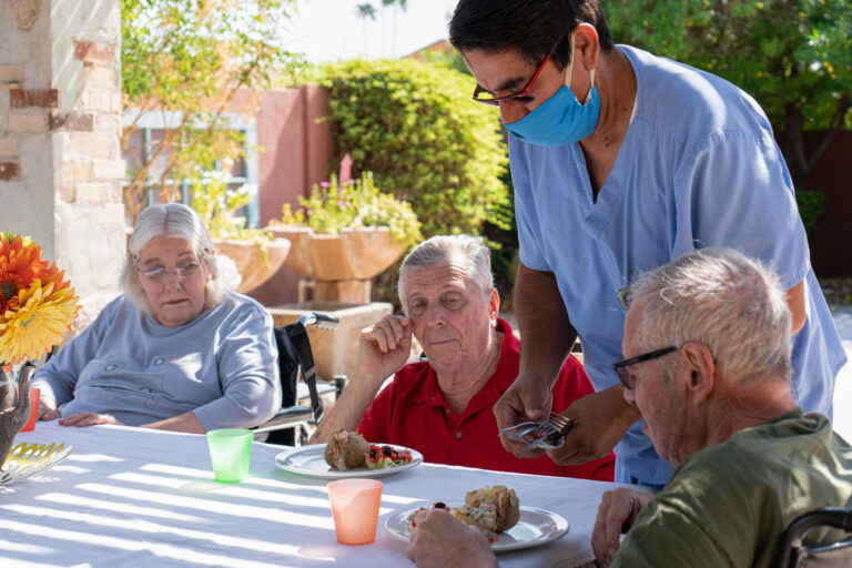 A group of seniors siting around the table and a caregiver distributing the spoon and fork.